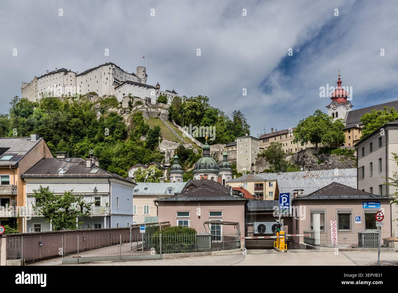 Blick von der Erzabt-Klotz-Straße auf die Festung Hohensalzburg und eine Kirche mit roter Kuppel; im Vordergrund der Eingang zu einem HYPO-Parkplatz. Die Festung Hohensalzburg (Baubeginn 1077) ist eine der größten und am besten erhaltenen mittelalterlichen Burganlagen Europas und überragt die Salzburger Altstadt auf dem Festungsberg (542 m ü. M.). Salzburg, Österreich Stockfoto