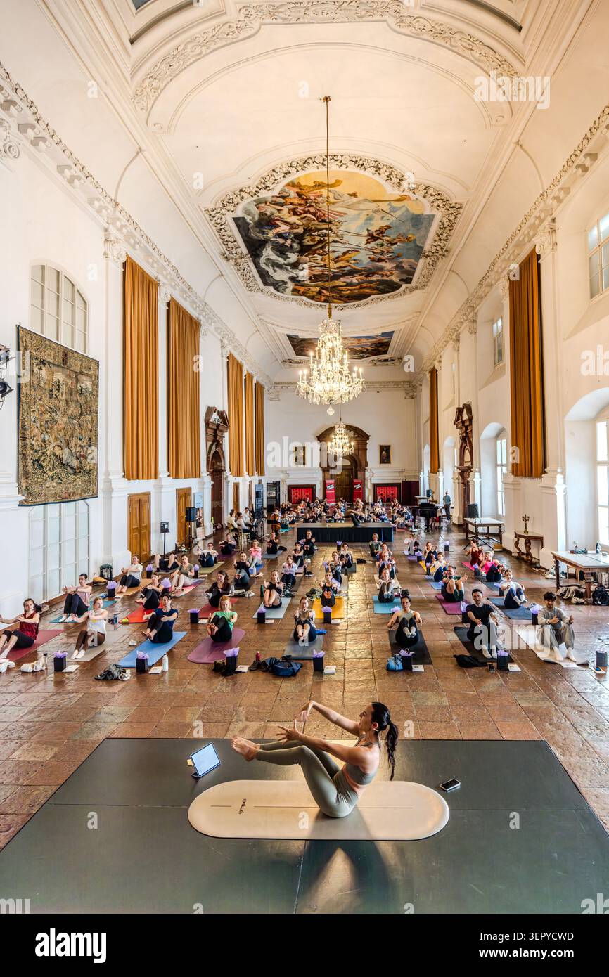 Blick auf den Ballsaal im DomQuartier Salzburg von der Galerie aus: Mehrere Dutzend Teilnehmer sitzen auf Matten; auf dem zentralen Podium führt ein Yogalehrer die Sitzung mit einem Kronleuchter und einem barocken Deckenfresko im Hintergrund. Veranstaltung „Pop Up Yoga – Mozart Edition“. Salzburg, Österreich Stockfoto