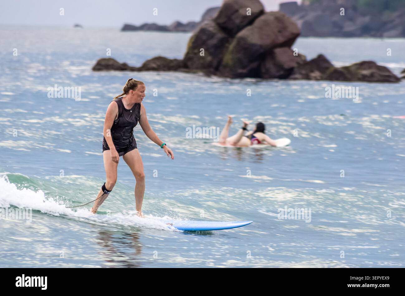 Mirissa, Sri Lanka - 20. Februar 2023: Eine Frau reitet auf einem blauen Longboard auf einer kleinen Welle, während ein anderer Surfer im Hintergrund in der Nähe von ihrem Board fällt Stockfoto