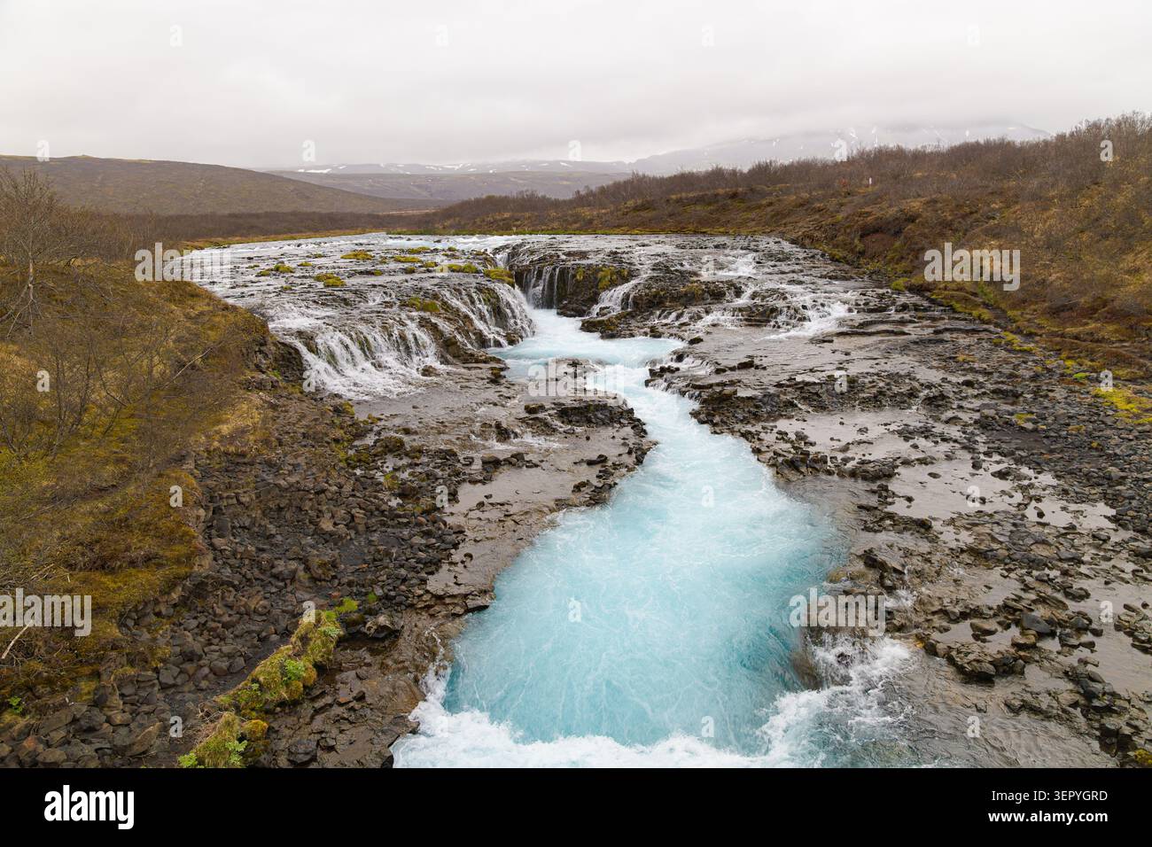 Der atemberaubende hellblaue Brúarfoss-Wasserfall, der in einen dunklen vulkanischen Felsschlucht unter einem bedeckten Himmel in Island stürzt. Stockfoto