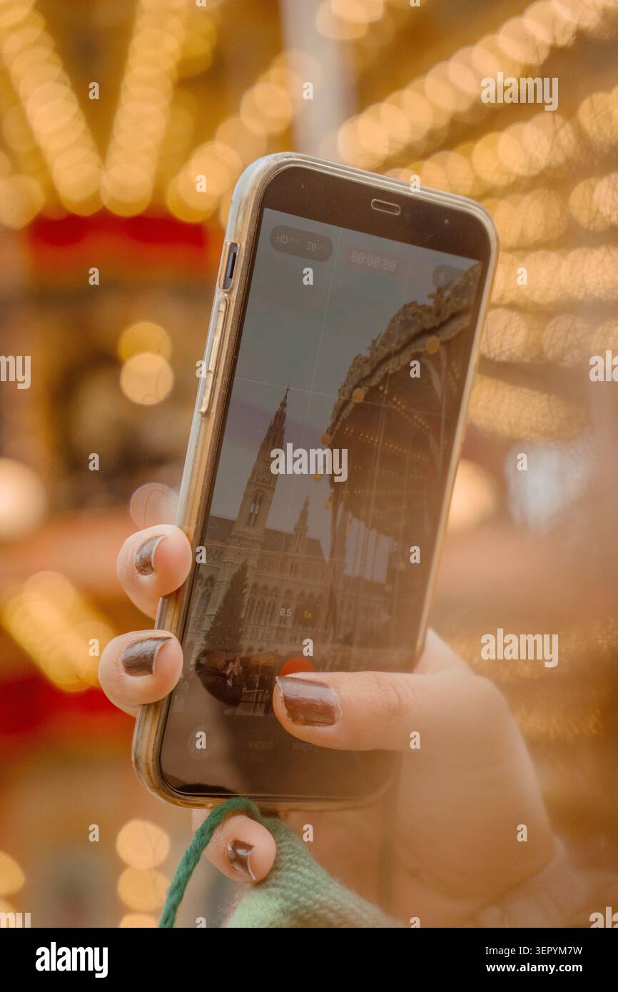Hand hält ein Smartphone, das das Rathaus und Karussell auf einem festlichen Weihnachtsmarkt bei Nacht erfasst. Vertikale Aufnahme. Stockfoto