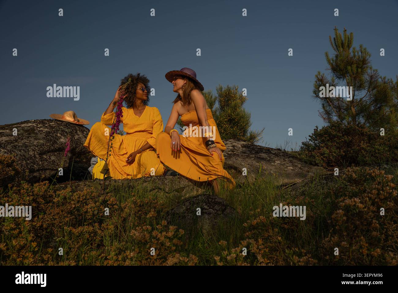 Zwei Frauen in gelben Kleidern sitzen bei Sonnenuntergang auf einem felsigen Hügel Stockfoto