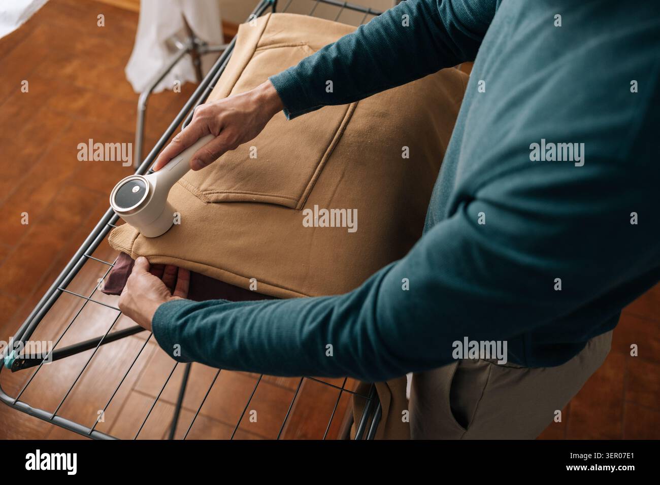 Hochwinkelaufnahme eines Mannes, der Flusen von einem beigen Sweatshirt mit einem modernen elektrischen Stoffrasierer entfernt, während er Hausarbeiten und Kleidungspflege durchführt Stockfoto