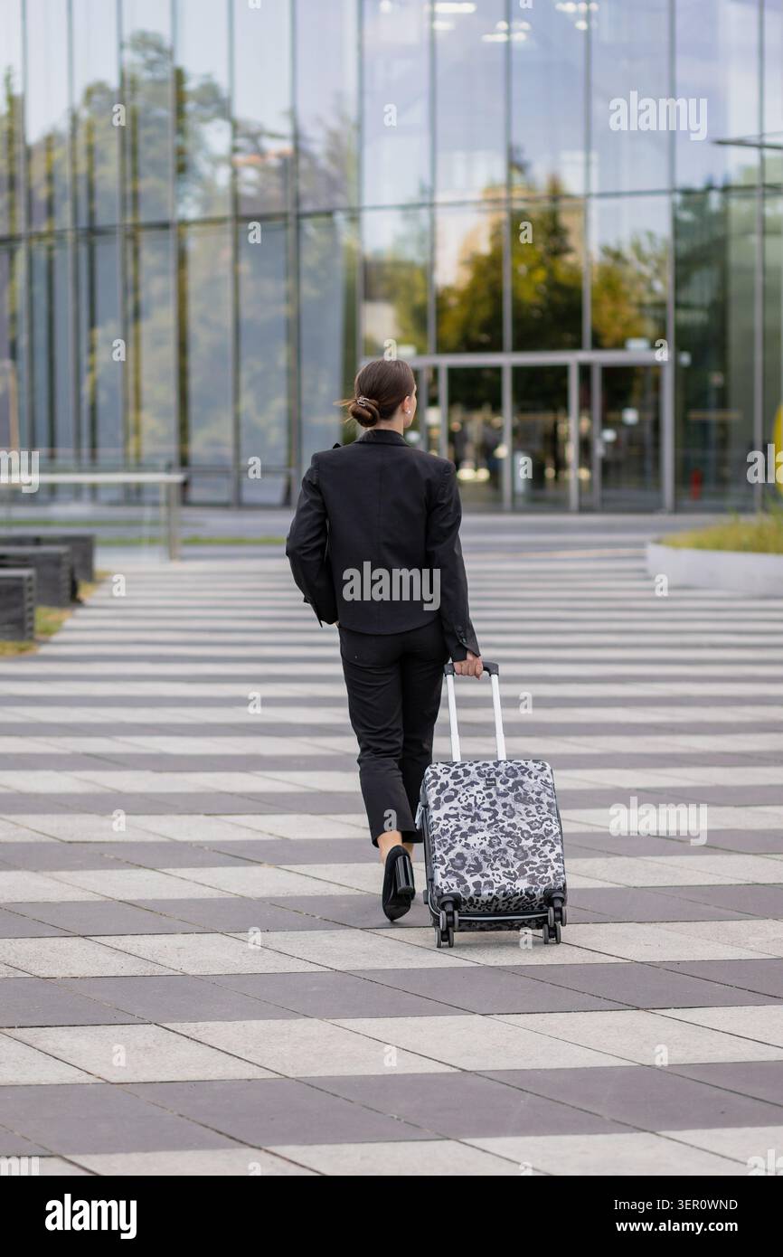Eine Person in einem schwarzen Anzug geht von der Kamera weg, während sie einen gemusterten Koffer zieht. Sie bewegen sich in Richtung eines großen Glasgebäudes in einer urbanen Umgebung Stockfoto