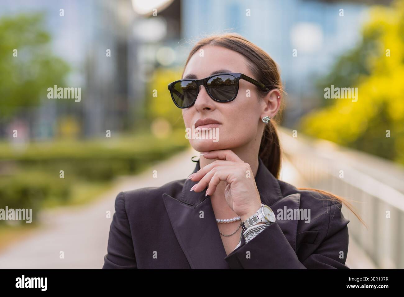 Eine Frau steht selbstbewusst in einem Stadtgebiet, trägt eine Sonnenbrille und eine schwarze Jacke. Sie hat ihre Hand am Kinn, blickt nach vorne, mit grünen Bäumen und Stockfoto
