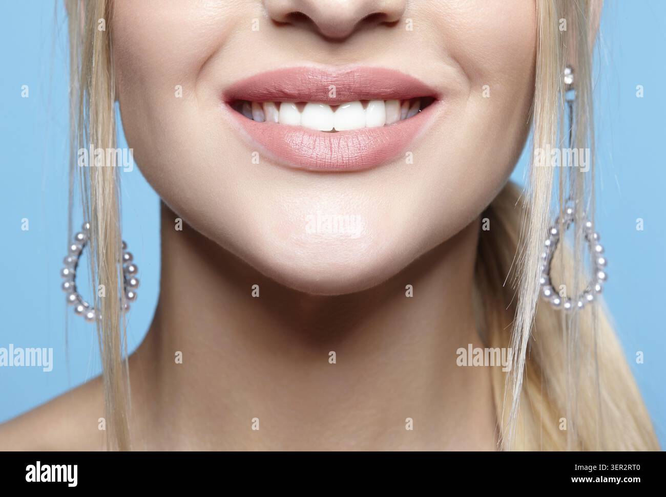 Makroporträt des weiblichen unteren Gesichtsbereichs. Blonge Frau mit roten, lächelnden Lippen, Make-up und Ohrringen. Mädchen auf blauem Hintergrund Stockfoto