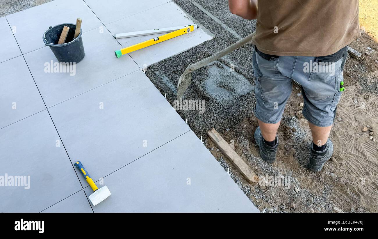 Männlicher Bauarbeiter, der Fliesen auf der Außenfläche legt, mit einer Kelle Klebstoff verteilt, wobei Werkzeuge und Materialien auf dem Boden sichtbar sind Stockfoto