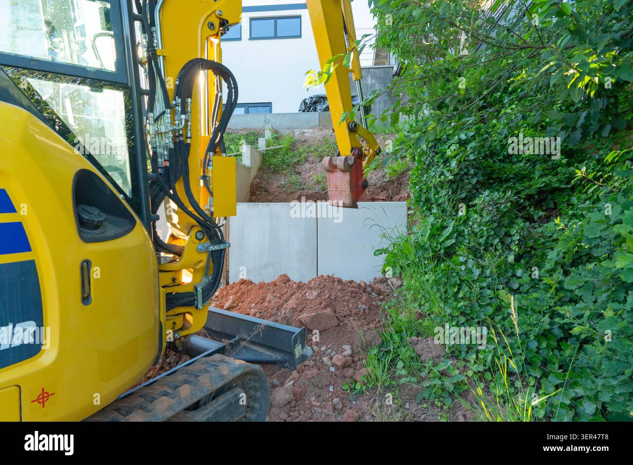 Ein gelber Minibagger wird während der Landschaftsgestaltung in einem Hinterhof eingesetzt und installiert Betonstützen in der Nähe eines grünen Hanges. Kompakt Stockfoto