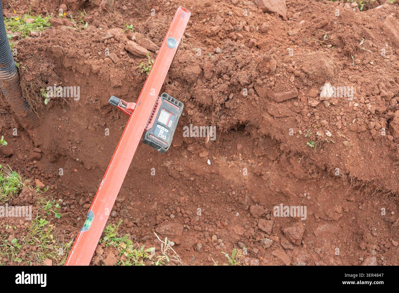 Ein professionelles orangefarbenes Wasserwaage-Messgerät mit digitalem Messempfänger, das in einem Schmutzgraben platziert ist, um die Genauigkeit der Neigung zu prüfen Stockfoto