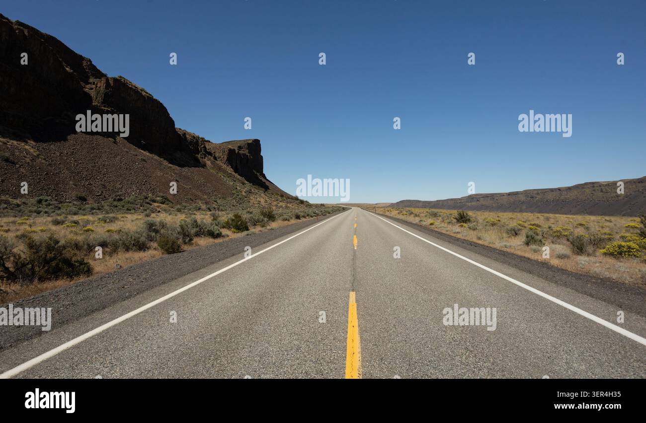 Ein gerader zweispuriger Highway erstreckt sich durch eine abgelegene Wüstenschlucht mit felsigen Basaltklippen und trockenem Buschland unter einem klaren blauen Himmel. Stockfoto