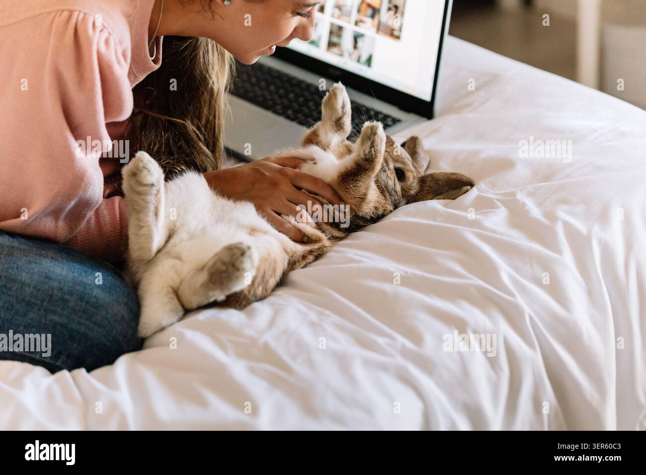 Frau, die ein Kaninchen auf einem weißen Bett streichelt, während sie einen Laptop benutzt Stockfoto