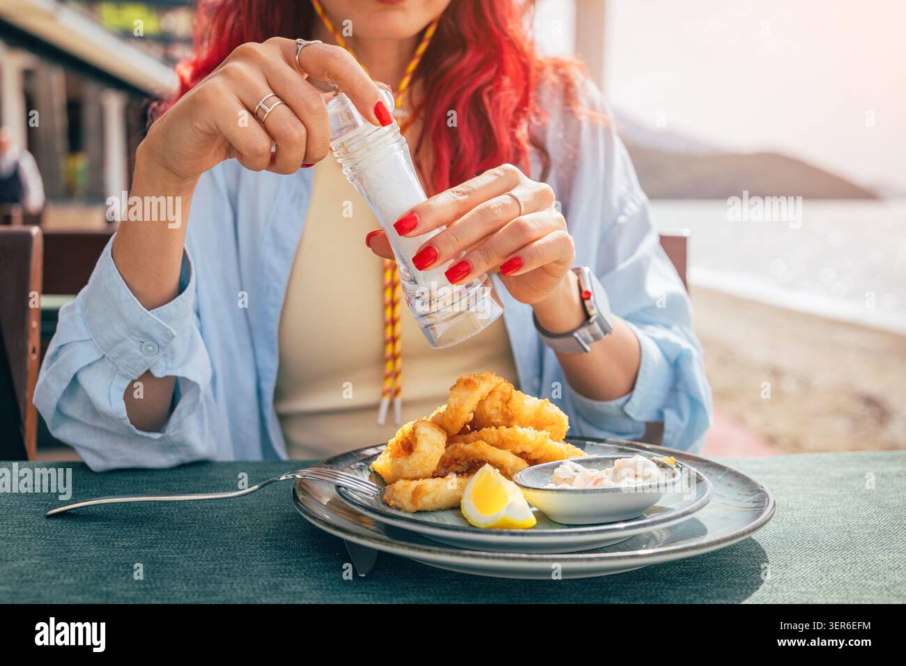 Frau, die Salz auf einen Teller knuspriger gebratener Calamari mit Zitrone und Soße gibt, und draußen am Meer isst Stockfoto