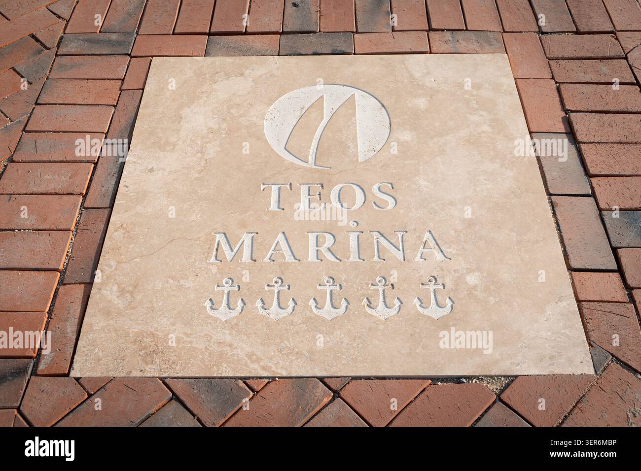 13. Mai 2026, Sigacik, Turkiye: Teos Marina Logo, Anker und Text in eine beige Steinplatte gehauen, umgeben von rötlichen Pflastersteinen Stockfoto