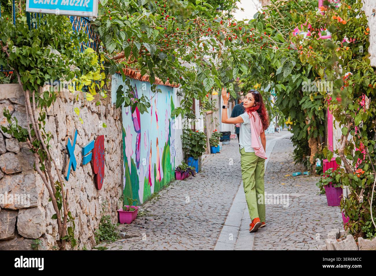 13. Mai 2026, Sigacik, Turkiye: Junge Frau, die entlang einer malerischen Kopfsteinpflasterstraße mit blühenden Pflanzen, Steinmauern und lebendiger Wandkunst spaziert Stockfoto