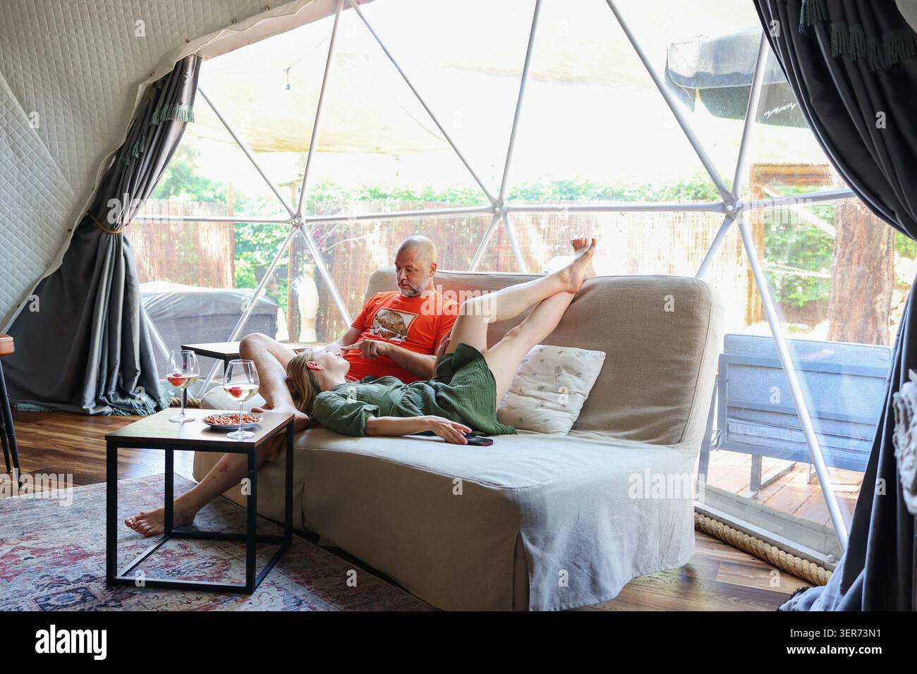 Gemütlicher Glamping-Lebensstil mit einem Paar, das drinnen in einer modernen geodätischen Kuppel inmitten der Natur ruht. Stockfoto