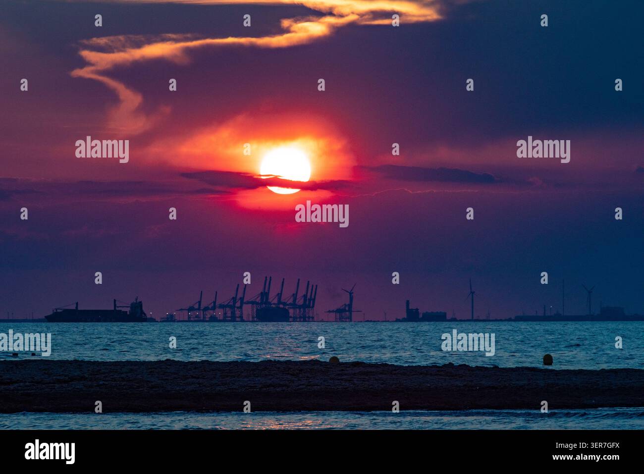Die Sonne untergeht orange und rot über einem Industriehafen, mit Containerkränen und Frachtschiff Stockfoto