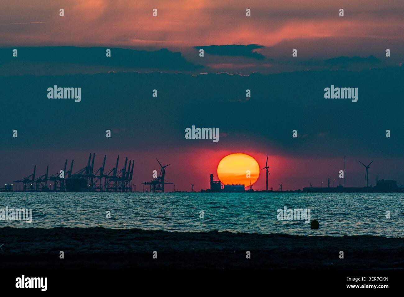Sonnenuntergang beleuchtet Windturbinen und Containerkräne an einem Industriehafen Stockfoto
