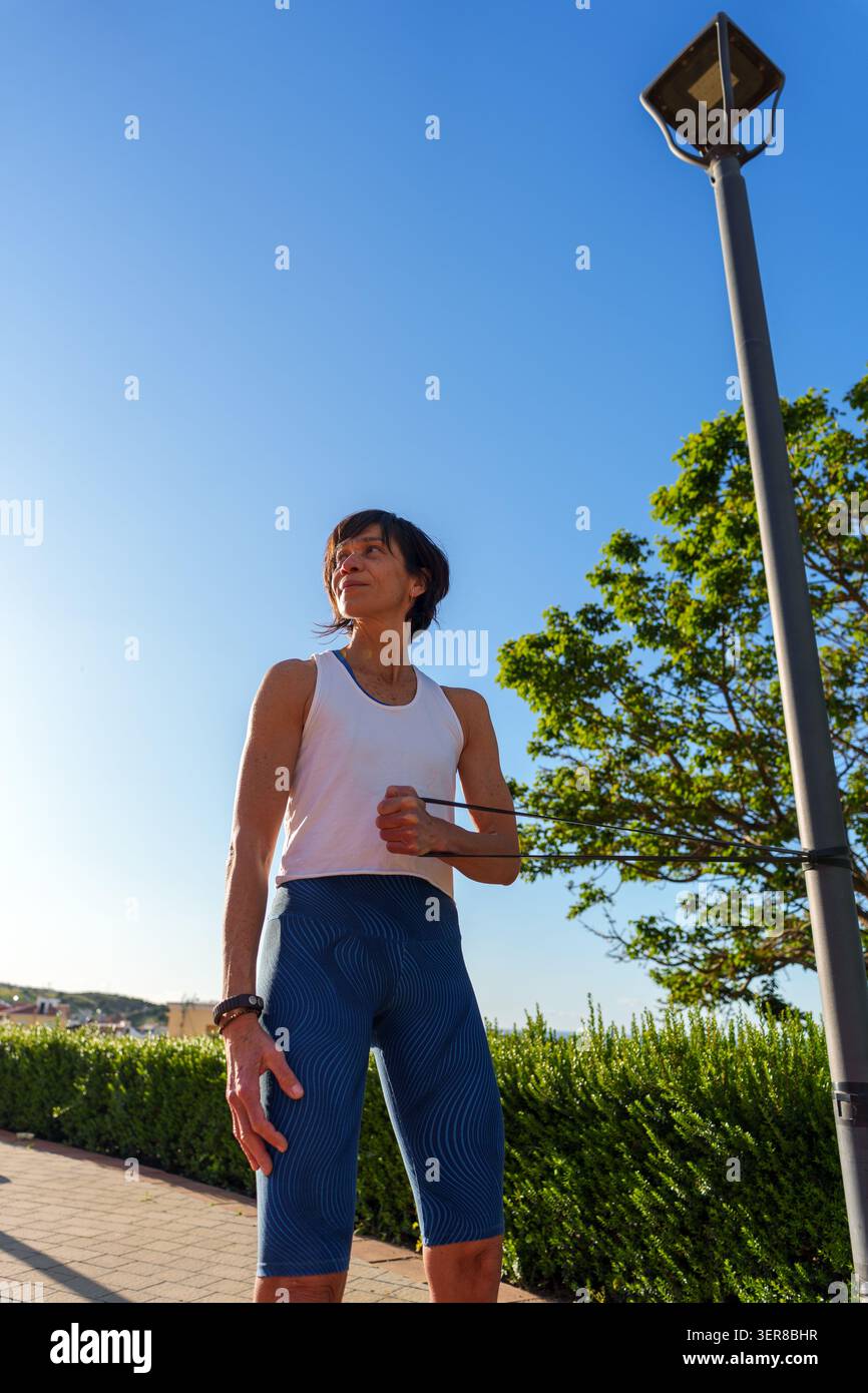 Reife Frau, die ein Widerstandsband-Workout durchführt, Arme und Schultern im Freien unter klarem blauen Himmel trainiert und einen aktiven Lebensstil fördert Stockfoto