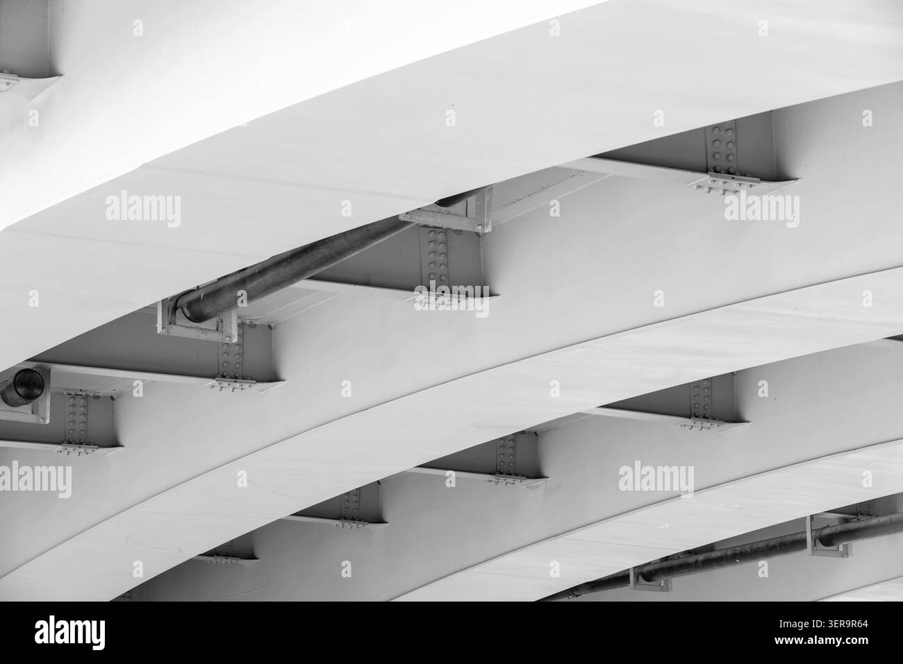 Unter einer modernen Stahlbrücke erzeugen sich wiederholende, geschwungene Stützbalken und genietete Bögen ein auffälliges monochromes Muster. Minimale architektonische Details Stockfoto