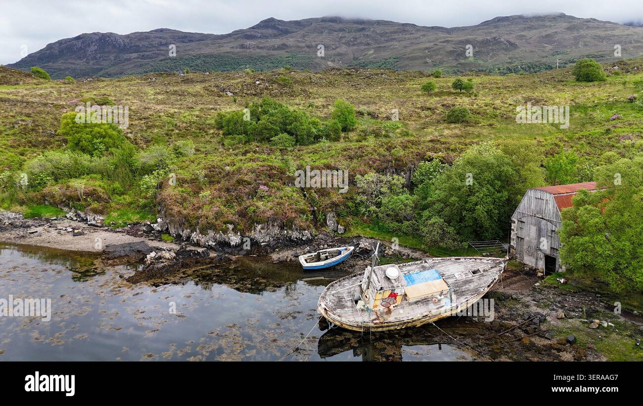 Eine ruhige Küstenlinie von Highland, an der verwitterte Boote, ein altes Bootshaus und zerklüftete Hügel die zeitlose Schönheit Schottlands einfangen Stockfoto