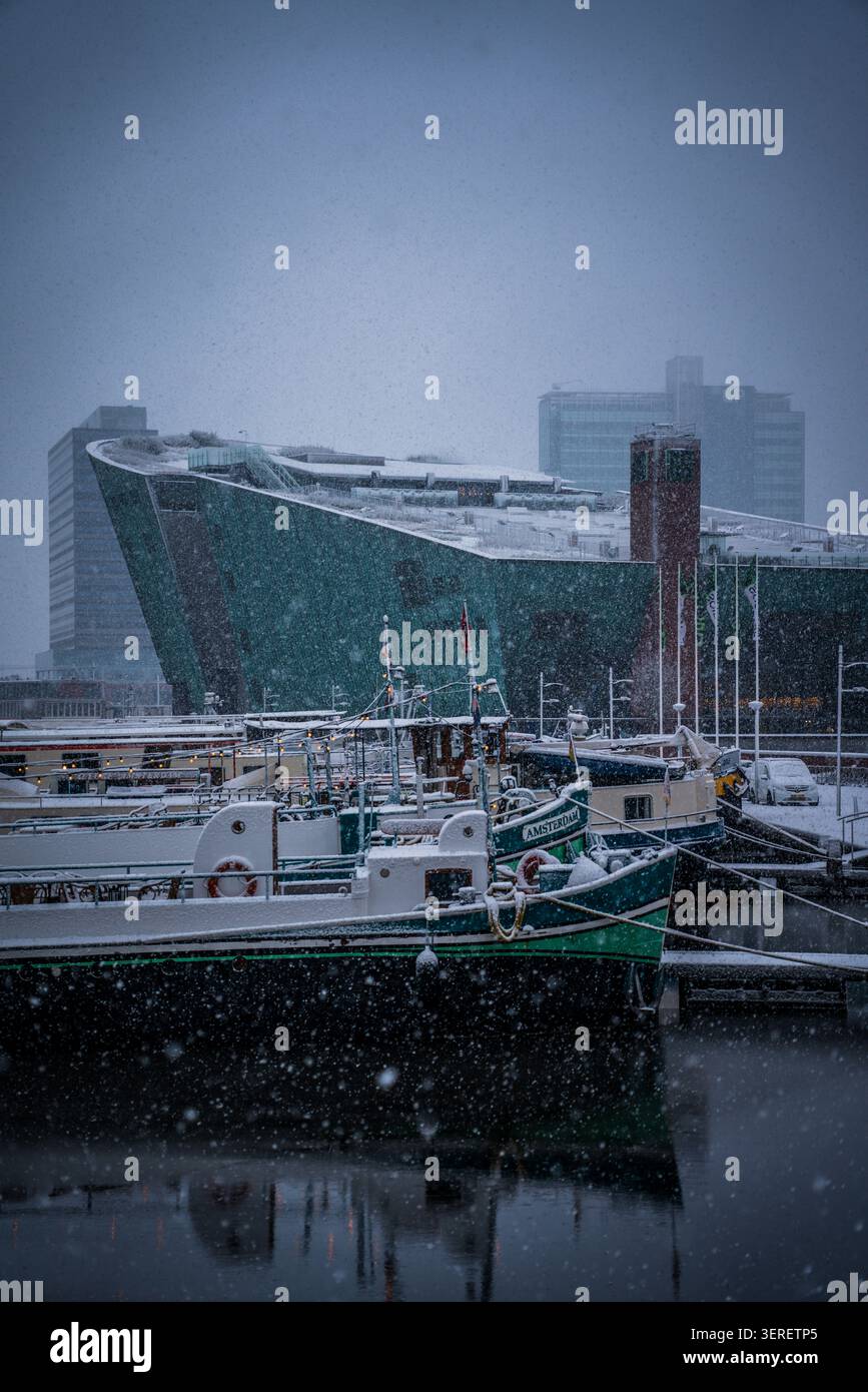 Eine friedliche Winterszene fängt den Schneefall über mehreren Booten ein, die in einem Hafen in Amsterdam, Niederlande, vertäut sind. Im Hintergrund ist die markante Grinse Stockfoto
