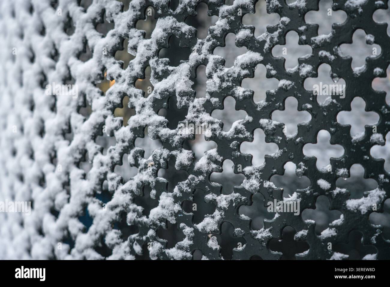 Bei einer Nahaufnahme wird frischer weißer Schnee auf einem dunklen, komplizierten Metallgitter mit dekorativen, quatrefoil-ähnlichen Mustern aufgenommen. Dieses atmosphärische Bild Stockfoto