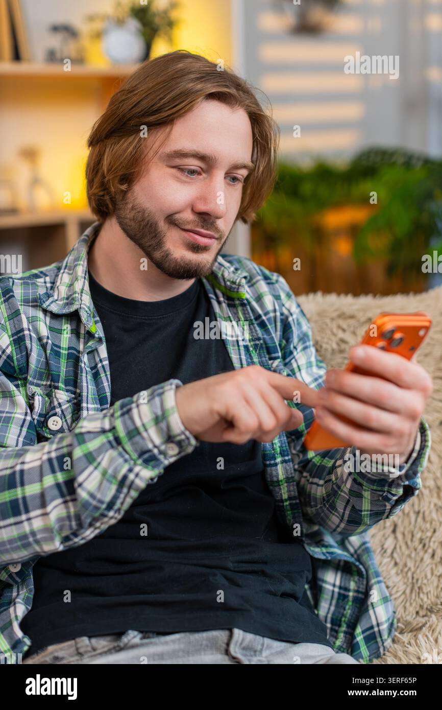 Ein junger Mann auf dem Heimsofa gibt eine schnelle Smartphone-Nachricht ein und hört dann auf, um Worte mit einem gezielten Lächeln erneut zu lesen. Erwachsener Kerl auf dem Heimsofa drückt mit ruhiger Zufriedenheit nach dem Lesen der SMS und dem Beenden des Chats senden. Stockfoto