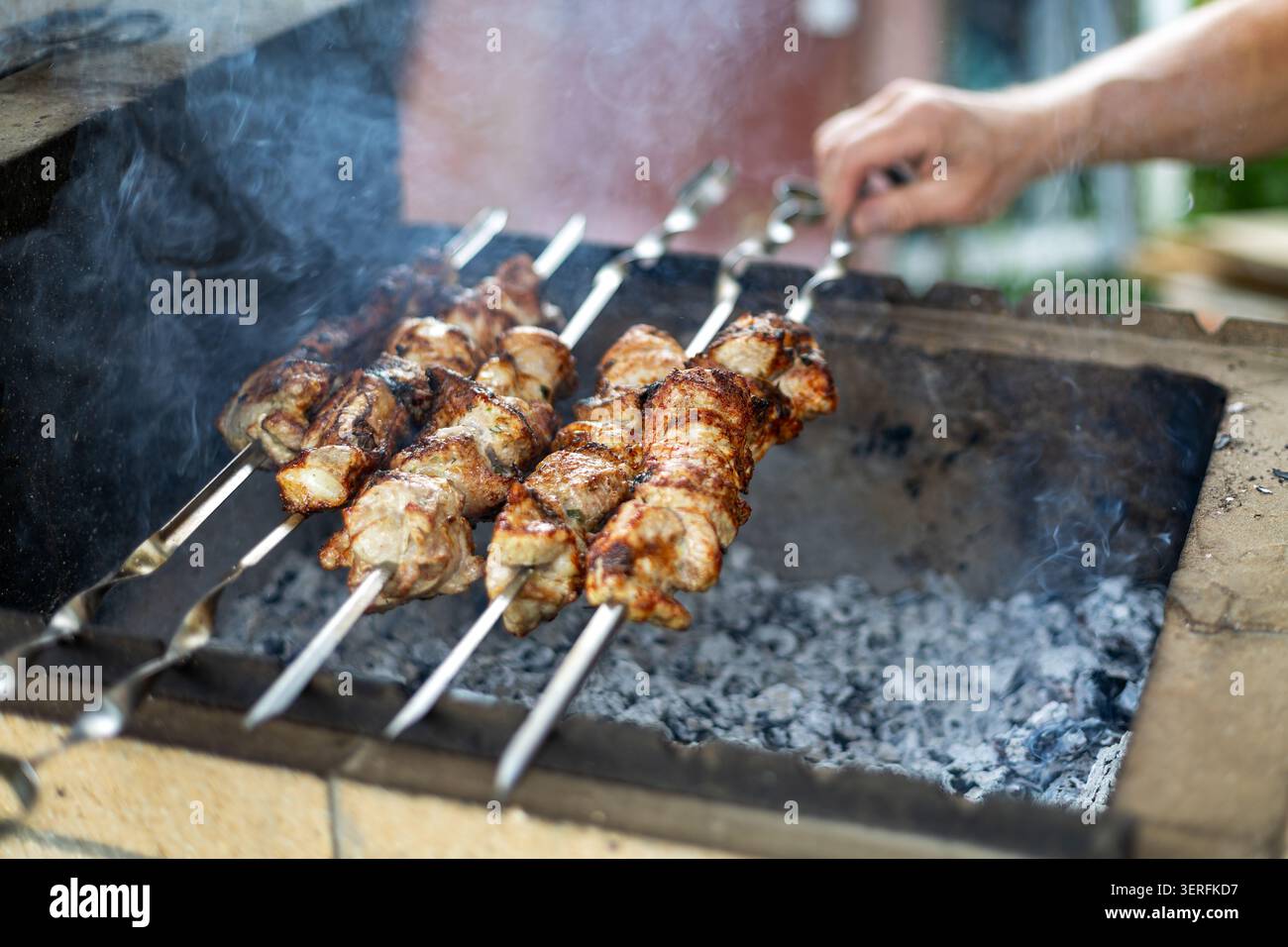 Gegrillte Fleischspieße, die über heißen Kohlen kochen. Gegrillte Fleischspieße kochen über rauchig heißen Kohlen, während eine Hand sie auf einem Grill im Freien dreht. Stockfoto