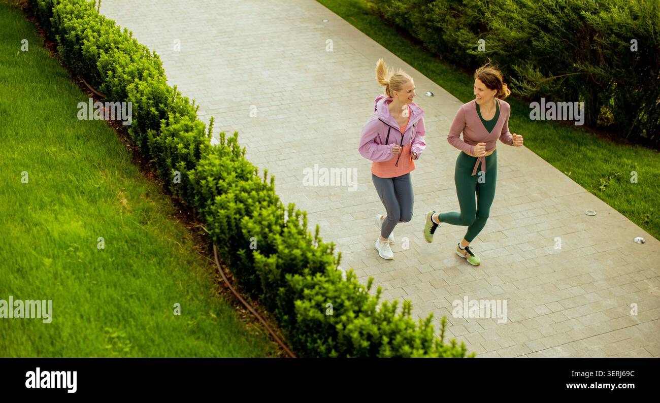 Zwei Frauen joggen an einem sonnigen Tag Seite an Seite auf einem glatten Weg. Sie genießen ihre Trainingsroutine, während sie von grünen Büschen und Gras umgeben sind. Stockfoto