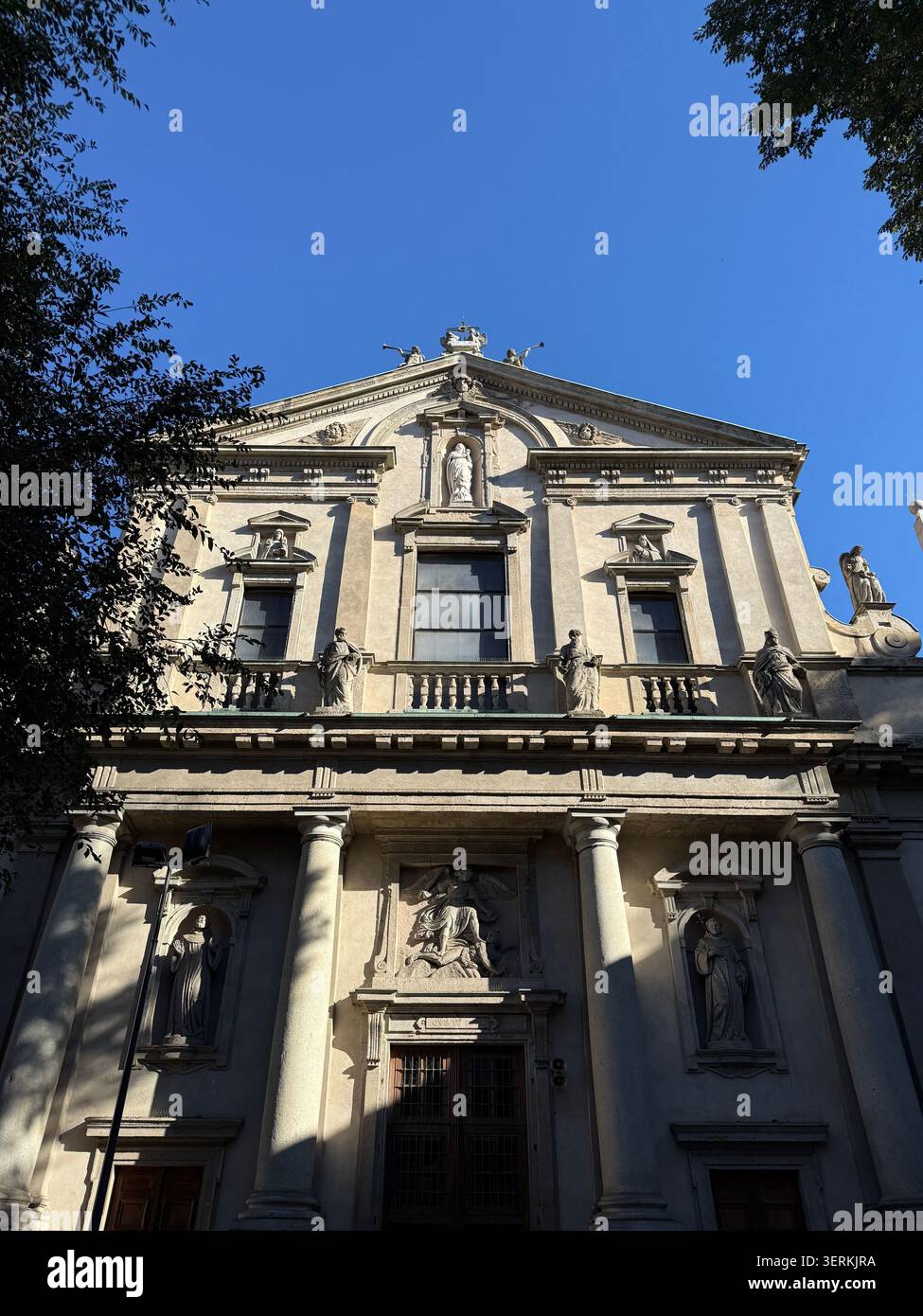 Mailand, Italien. Oktober 2025. Kunstvoll verzierte barocke Steinfassade einer historischen italienischen Kirche, die von Grün unter einem klaren blauen Himmel eingerahmt ist Stockfoto