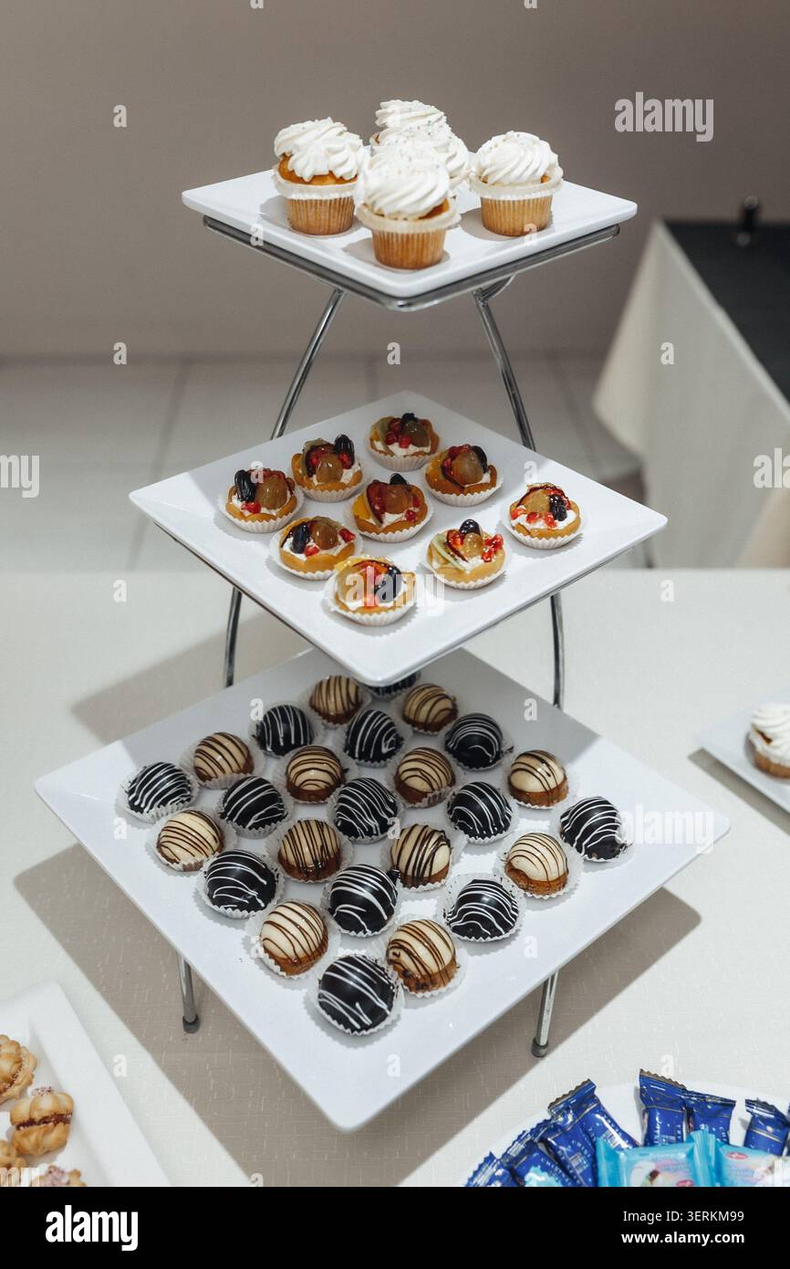 Der dreistufige Ständer bietet Platz für verschiedene Gebäckstücke. Weiße Teller zeigen Cupcakes, Torten und mit Schokolade überzogene Leckereien. Schlanke, silberne Beine unterstützen das elegante dess Stockfoto