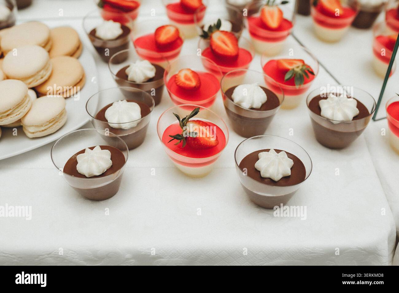 Süße Desserts glänzen unter weichem Licht. Cupcakes, Mousse und Macarons zieren eine weiße Tischdecke. Rote Erdbeergarnitur sorgt für hellen Kontrast. Schlagseite c Stockfoto