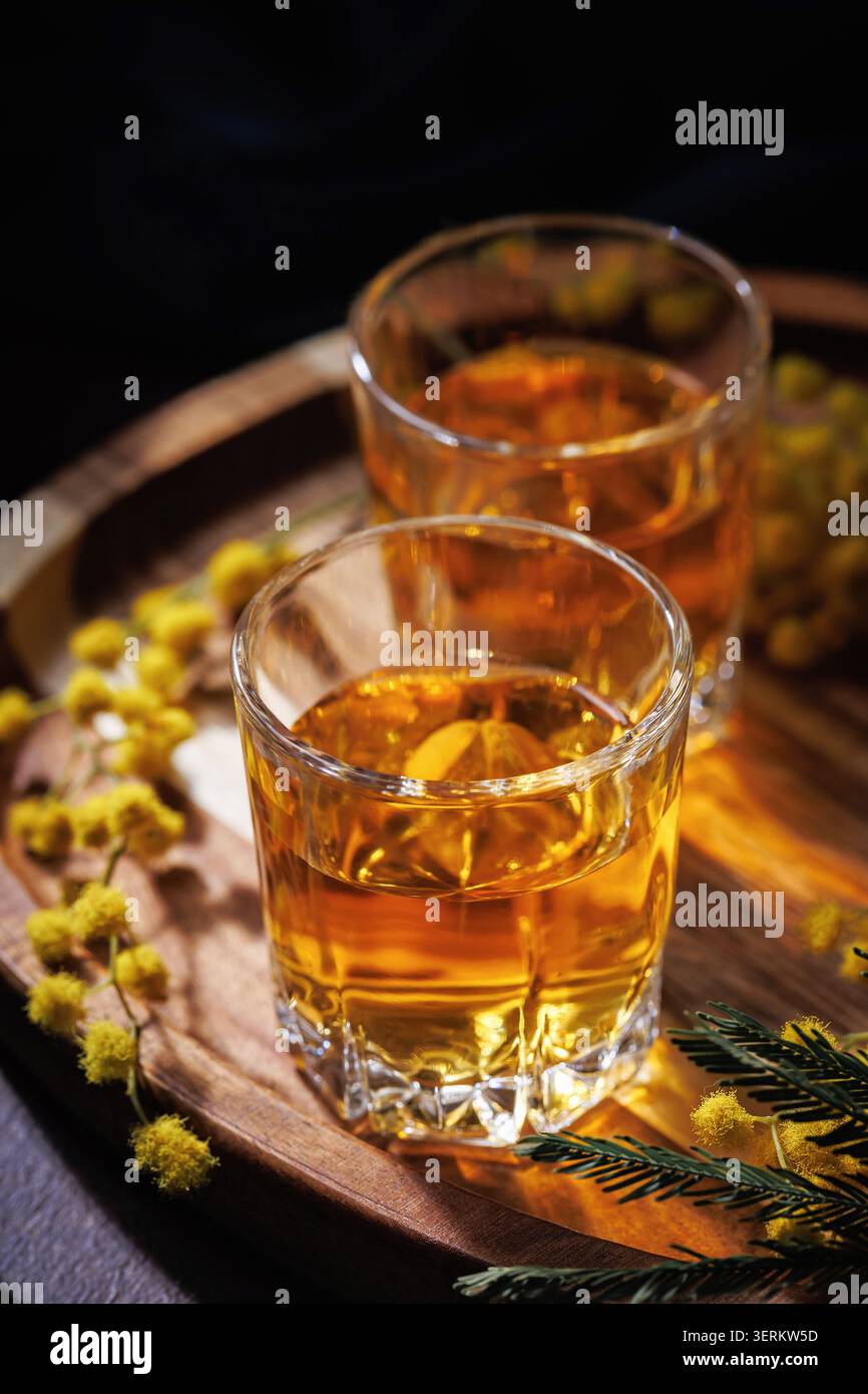 Whiskey- oder Cognac-Gläser mit Mimosa-Blumen auf Holztablett elegante Frühlingsalkoholszene Stockfoto