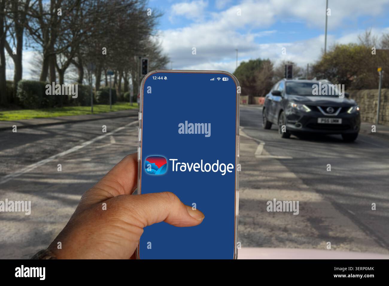 Die Hand hält ein Smartphone mit Travelodge Logo und App auf einem Bildschirm, mit einem Nissan Qashqai auf einer britischen Straße im Hintergrund Stockfoto