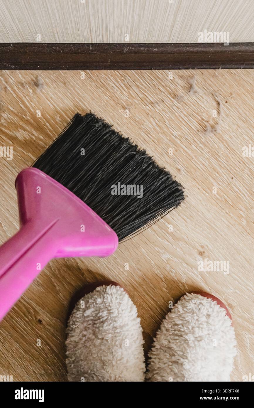 Rosafarbene Kunststoffbürste mit schwarzen Borsten, die Staubrückstände von hellem Holzlaminatboden fegen, weiße, flauschige Hausschuhe unten sichtbar, oben nach unten Stockfoto
