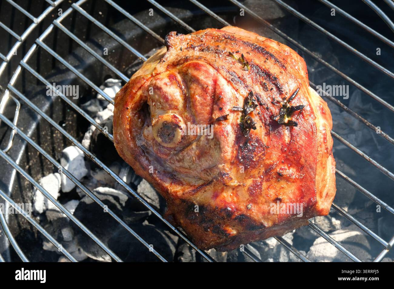 Ein Knochen-in-Schulter-Gelenk von Lammbraten auf einem Holzkohlegrill im Freien, gewürzt mit frischem Rosmarin und Kräutern während des Kochprozesses. Stockfoto