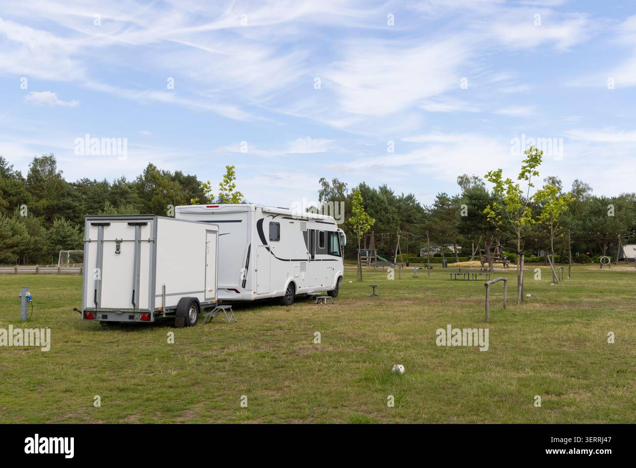 Ein weißes Wohnmobil und ein angehängter Anhänger auf einem ruhigen grasbewachsenen Campingplatz, umgeben von Bäumen und einem kleinen Spielplatz Stockfoto