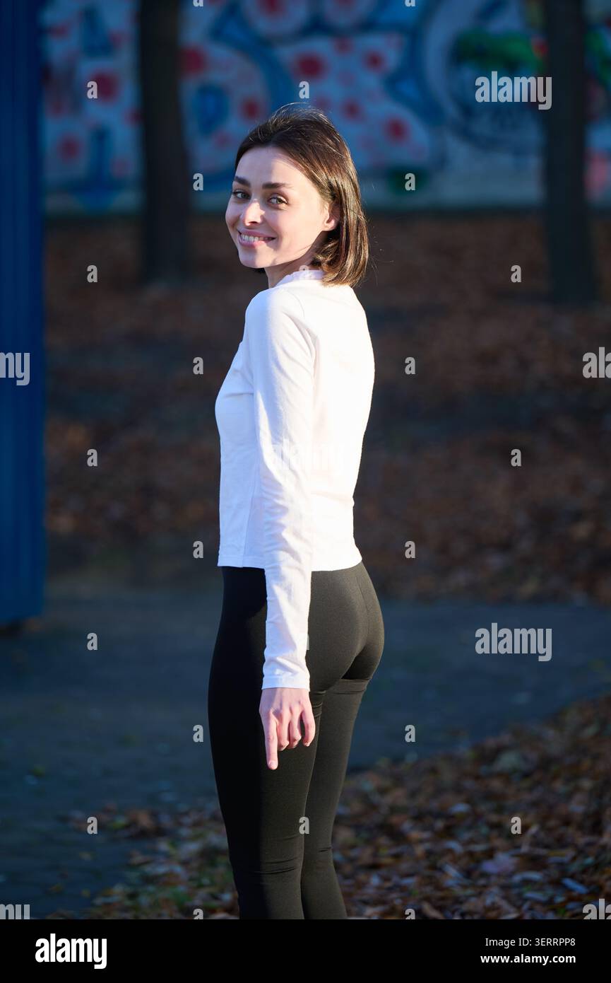 Junge Frau steht draußen und lächelt zurück in die Kamera, Rückansicht in weißem Oberteil und schwarzer Leggings. Nützlich für aktiven Lebensstil, Wellness, Freizeitmode oder Jugendkampagnen Stockfoto