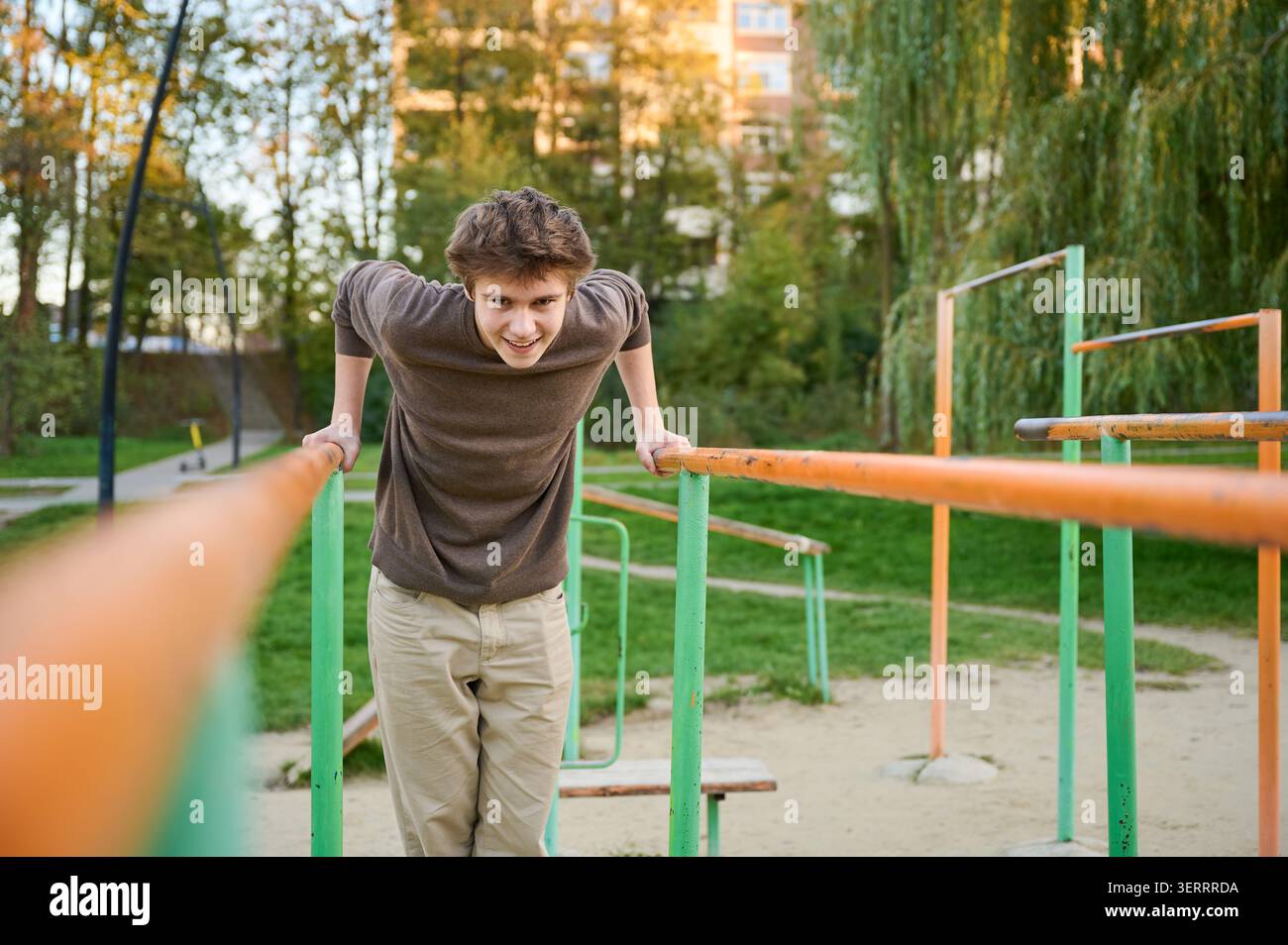 Weißer athletischer Mann, der parallele Dips mit kontrollierter Haltung durchführt, Calisthenics-Park-Umgebung im Freien mit orangegrüner Ausrüstung, intensivem Blick, Wohnblöcken und Bäumen im Hintergrund, Stockfoto