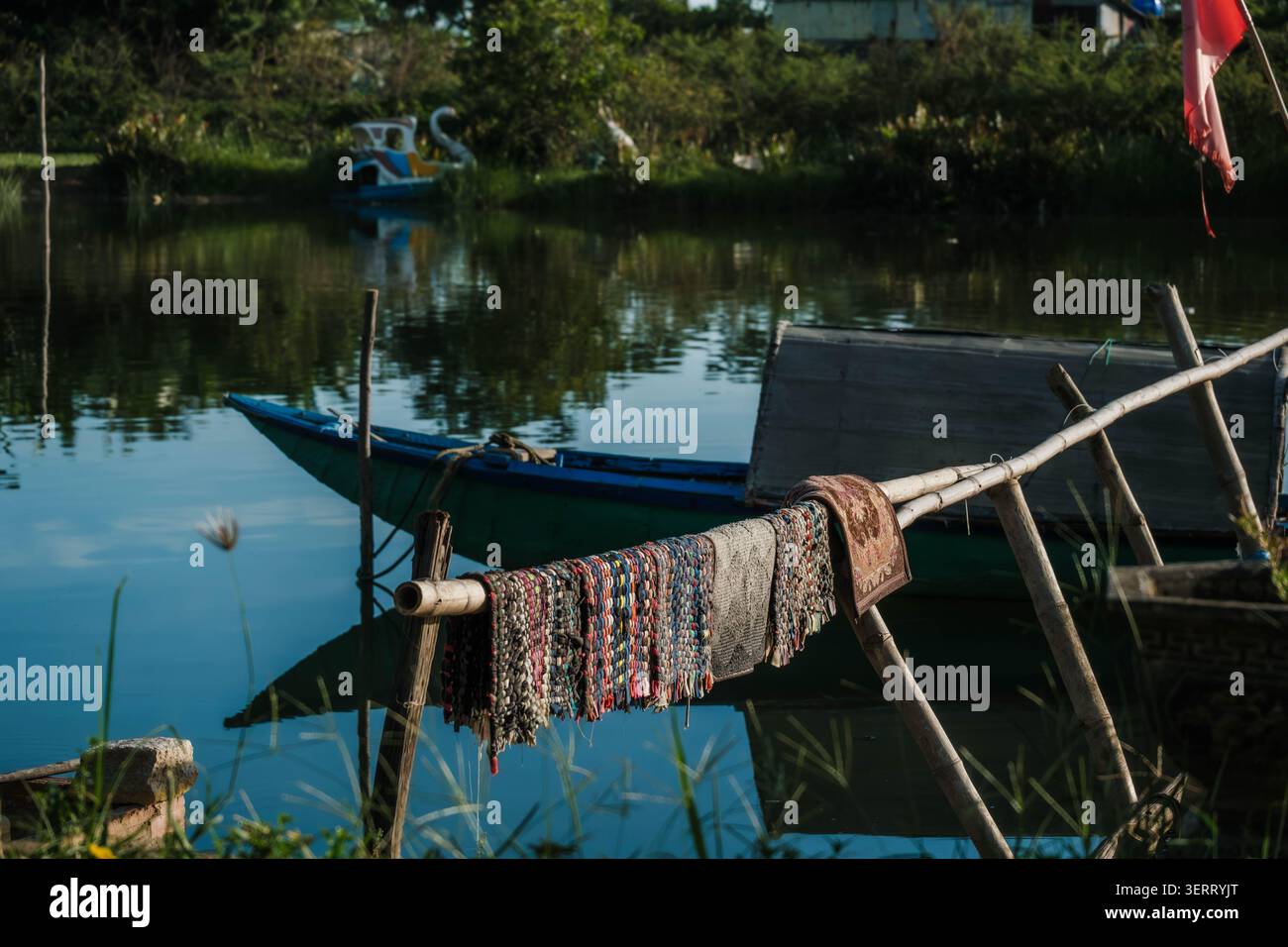 Gewebte Matten trocknen auf einem Bambusständer neben einem blauen Holzboot auf einem Fluss Stockfoto