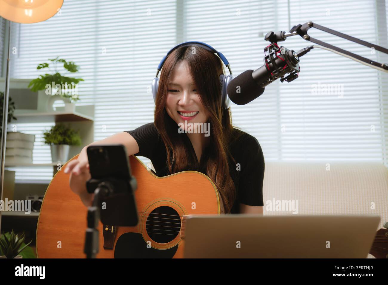 Asiatische junge Frau spielt Akustikgitarre im Heimstudio. Sie lächelt und zeigt während des Live-Streamings ein glückliches Engagement. Ihr Smartphone zeichnet Leistung auf Stockfoto