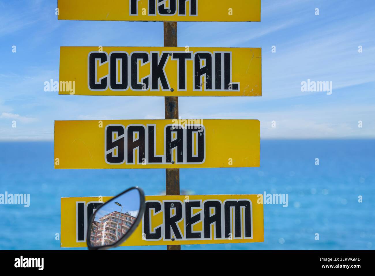 Auf dem gelben Menüschild stehen Speisen wie Fisch, Cocktails, Salat und Eis, wobei das Meer und der Himmel im Hintergrund sichtbar sind Stockfoto