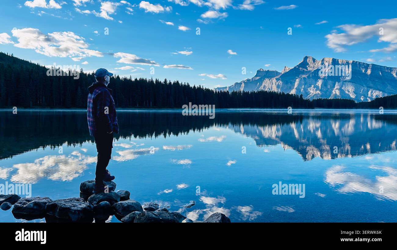 Silhouette einer Person, die am felsigen Ufer des Two Jack Lake steht und die perfekte Spiegelreflektion des Mount Rundle bei Sonnenaufgang, Banff National, bewundert Stockfoto