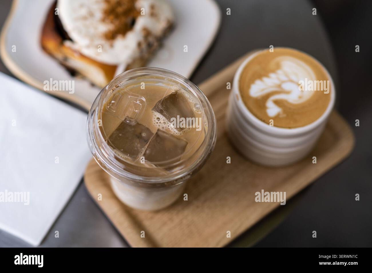 Eine gemütliche Café-Szene mit einem Latte mit komplizierten Schaumkunstwerken, einem Eiskaffee und einem überzogenen Dessert, alles auf einem Holztablett angeordnet. Stockfoto