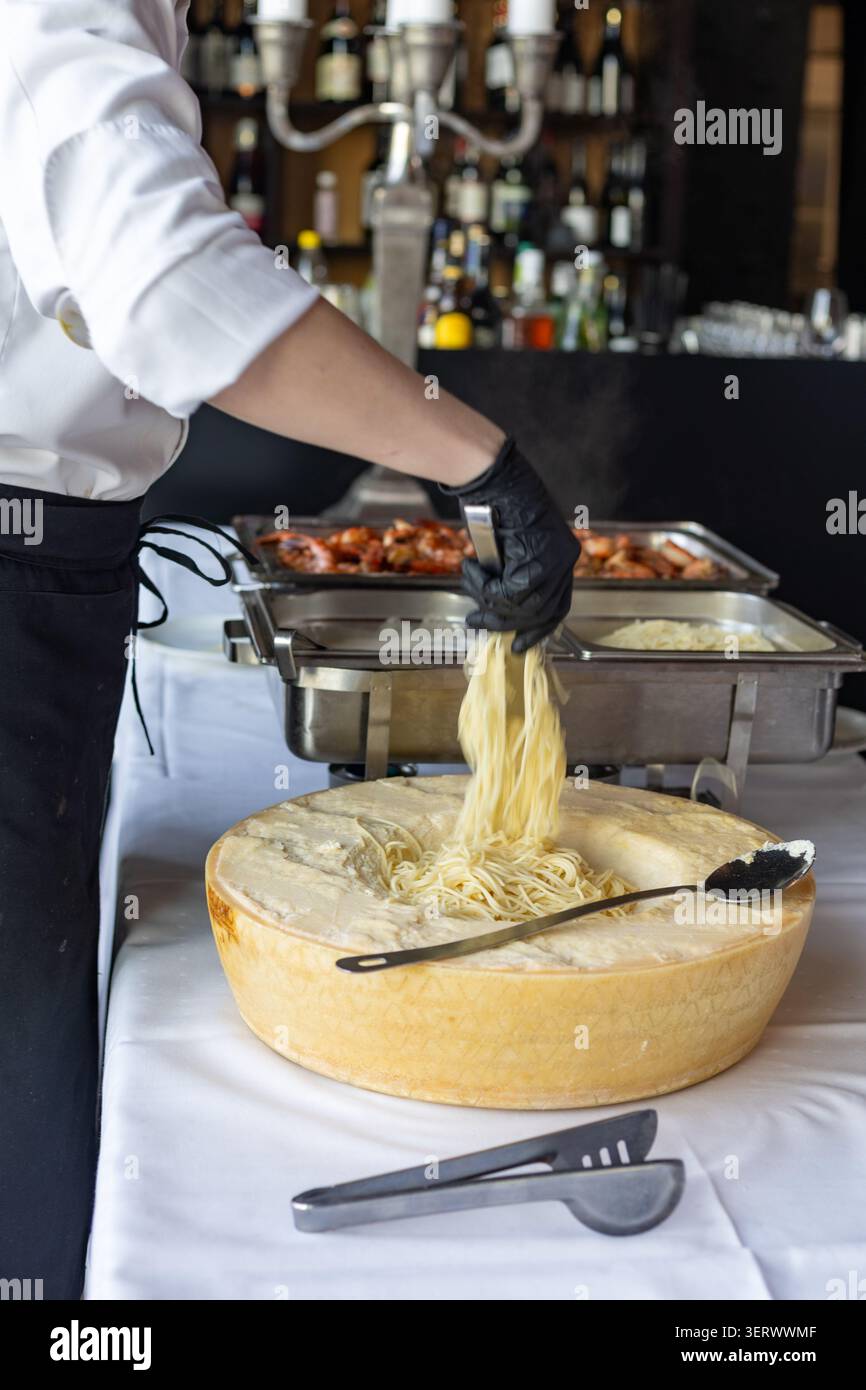 Ein Koch in einem schwarzen Handschuh zieht Nudelstränge aus einem großen Stück Käse. Die Küche ist aktiv mit Essenstabletts und Dampf in der Luft. Kunden wai Stockfoto