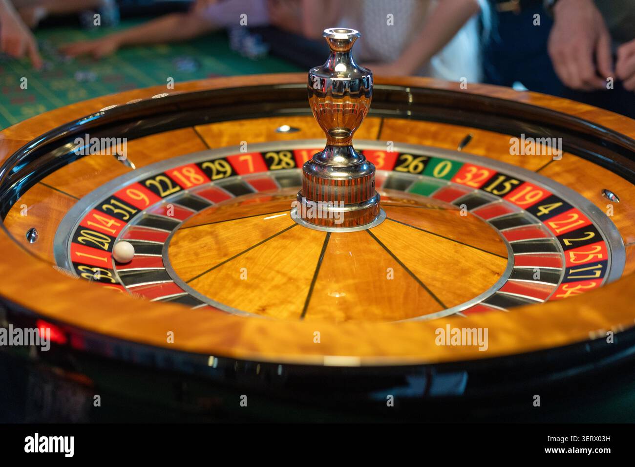 Eine Gruppe von Leuten versammelt sich um den Roulette-Tisch. Sie legen abwechselnd Wetten ab, während sich der Croupier darauf vorbereitet, das Rad zu drehen. Die Atmosphäre ist lebhaft Stockfoto