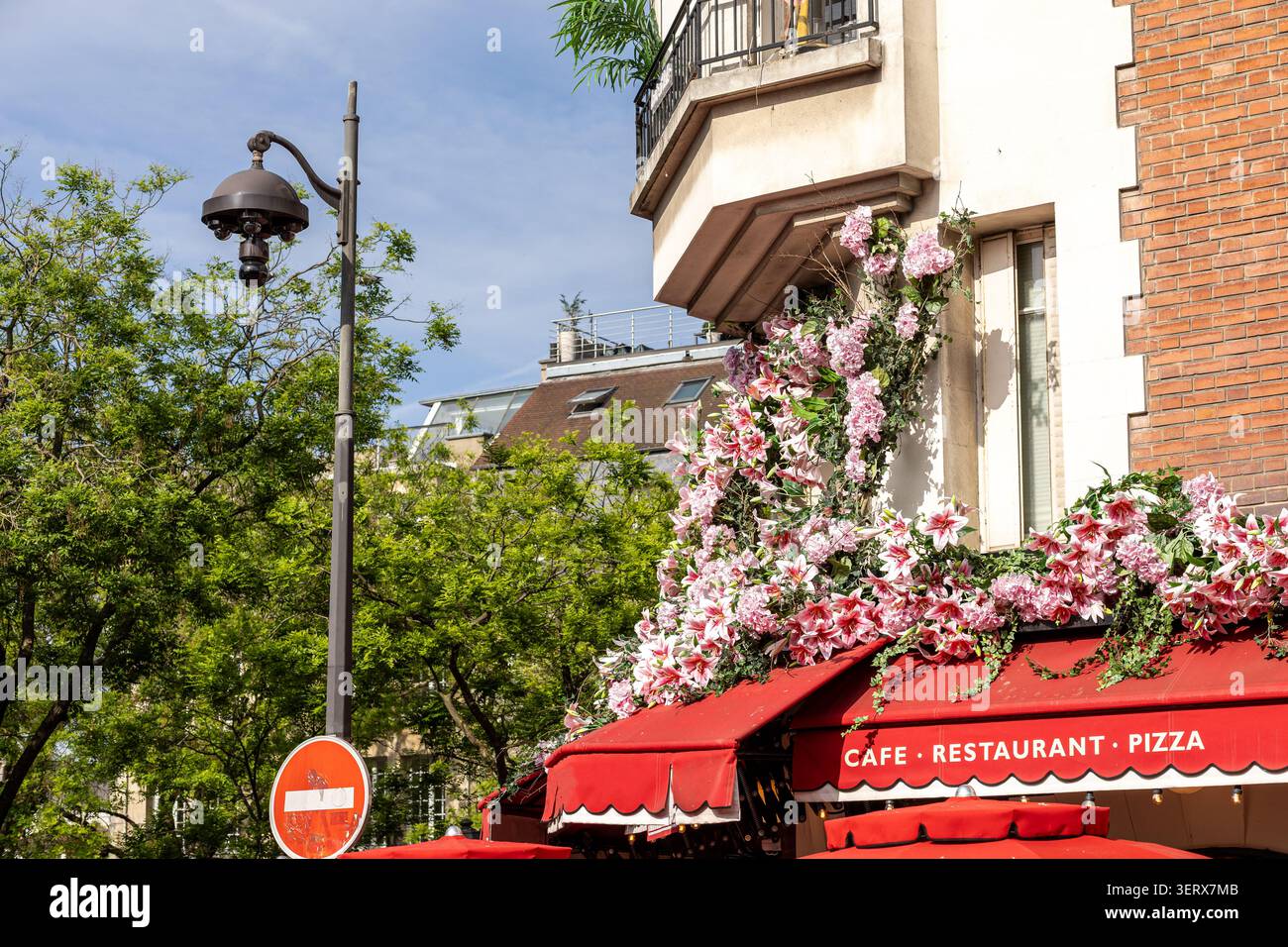 Helle Blumen bedecken den Außenbereich eines Cafés und Restaurants in städtischer Umgebung. Es ist ein sonniger Tag mit klarem Himmel und grünen Bäumen in der Nähe. Menschen Stockfoto