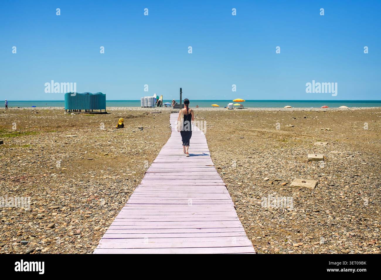 Batumi, Georgia – 18. Juni 2026: Malerischer Blick auf einen Kieselstrand mit einer langen lila Holzpromenade und einer jungen Frau, die zum Meer geht. Stockfoto