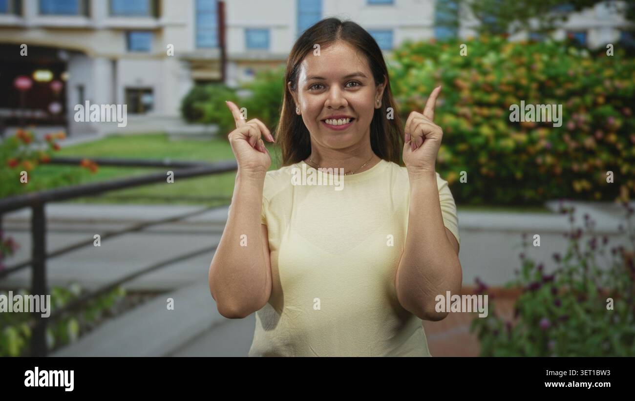 Die Frau zeigt beide Zeigefinger nach oben vor den Eingang des Gebäudes und lächelt in die Kamera; Optimismus wird ermutigt. Stockfoto