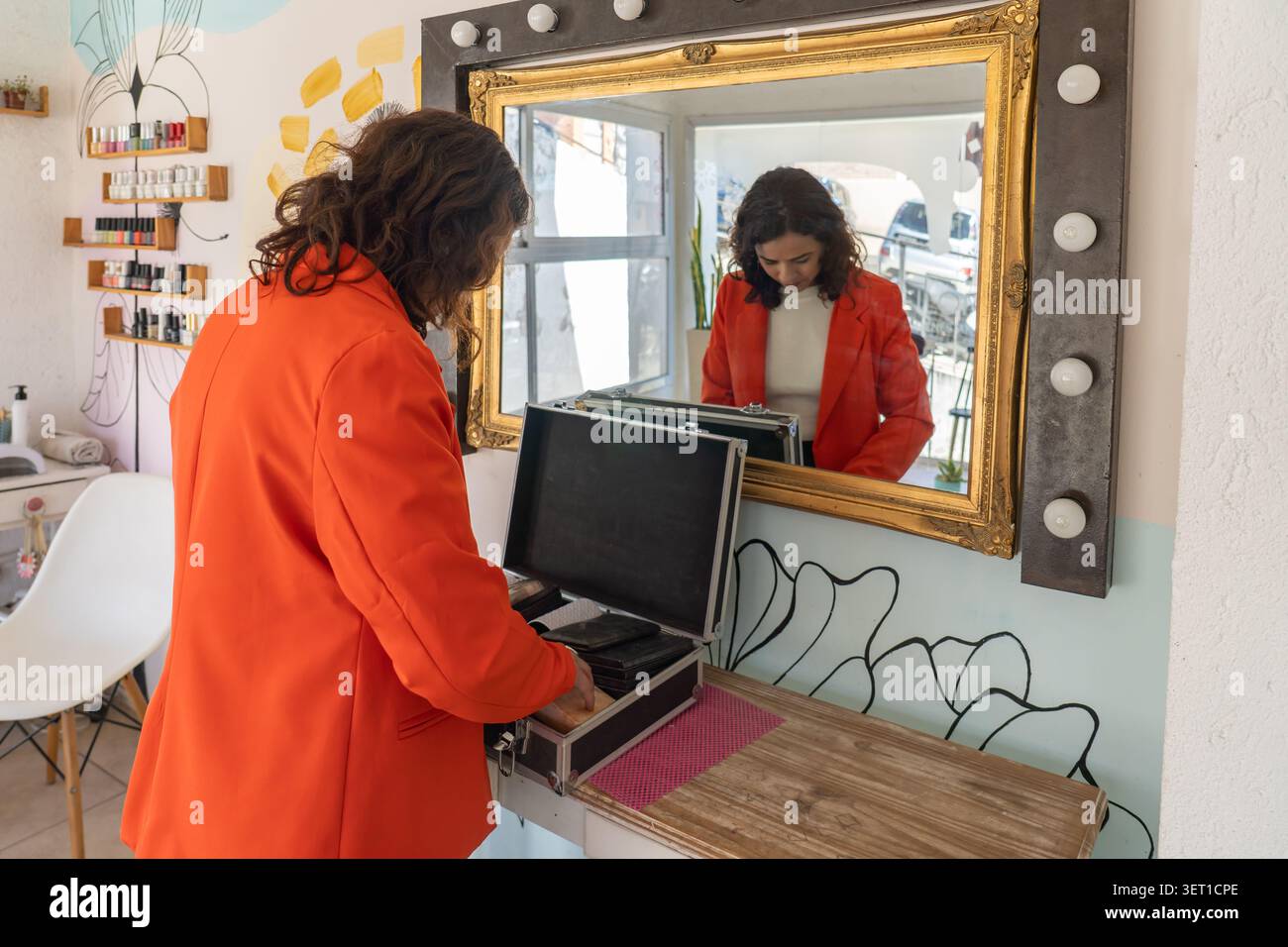 Frau in einem orangefarbenen Blazer, die eine professionelle Schminkhülle auf einem Kosmetiktisch neben einem Hollywood-Spiegel in einem Schönheitssalon öffnet Stockfoto